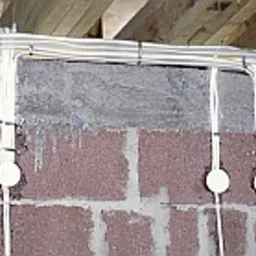 Pionowe i poziome kable elektryczne prowadzone wzdłuż szarej ściany z bloczków, przymocowane opaskami zaciskowymi, z okrągłymi puszkami instalacyjnymi w kolorze białym.